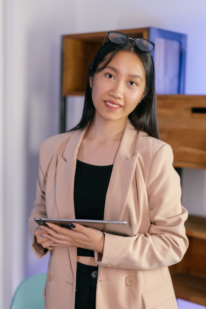 Asian businesswoman holding a tablet, wearing a beige blazer and sunglasses indoors.