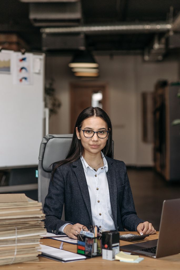 Asian businesswoman wearing glasses and suit, working at her desk in a contemporary office.