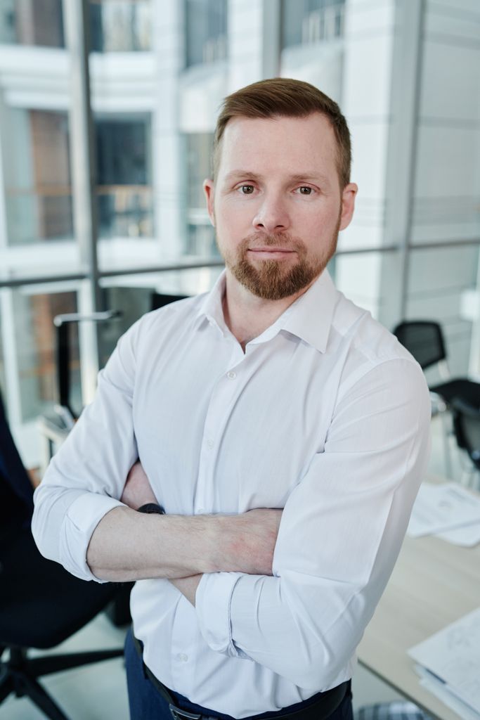 Confident man in white shirt posing in a modern office environment, standing with arms crossed.
