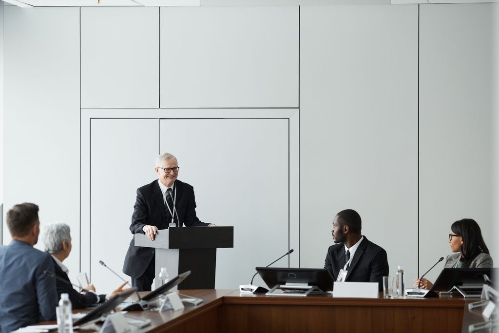 Formal business meeting with a diverse group and speaker at a podium in a conference room.
